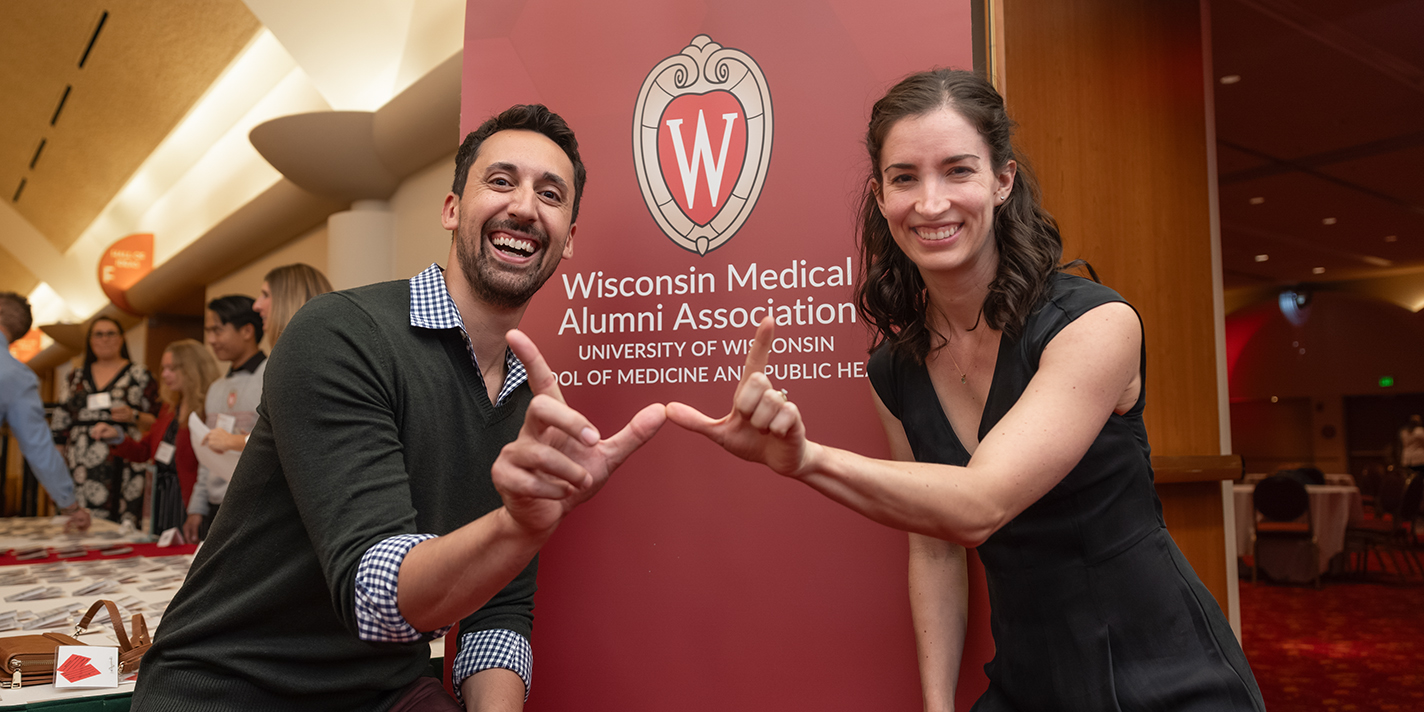 Two people join hands in forming the 'W' in front of a sign that reads 'Wisconsin Medical Alumni Association'